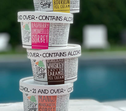 Stack of four colorful alcohol-infused ice cream pints (raspberry limoncello, chocolate whiskey salted caramel, mango margarita, bourbon) stacked poolside with a blurred backyard pool in the background; lids read contains alcohol, 21 and over.