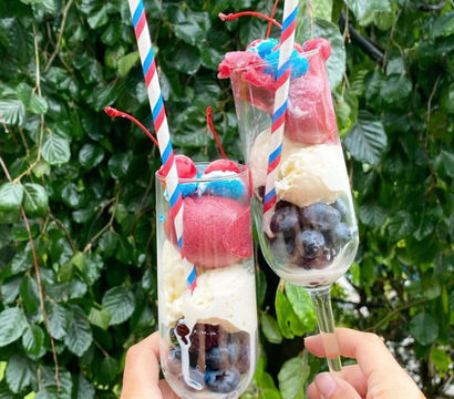 Two summer dessert parfaits in tall champagne flutes with blueberries, vanilla ice cream, red berry sorbet, cherries and red-white-blue striped paper straws held outdoors against leafy green foliage