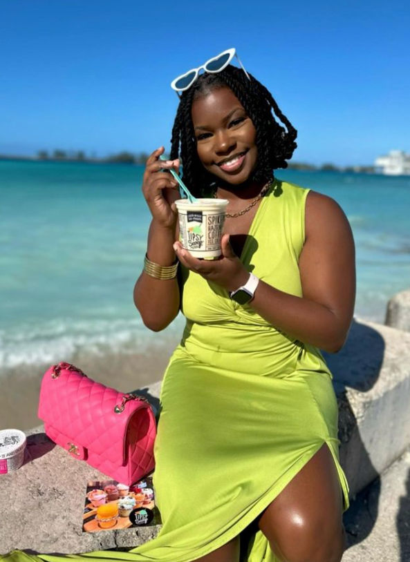 Smiling woman in a lime-green dress sitting on a seawall at a tropical beach with clear blue water and sky, white sunglasses on her head, enjoying a frozen treat from a cup with a spoon, bright pink quilted purse beside her.