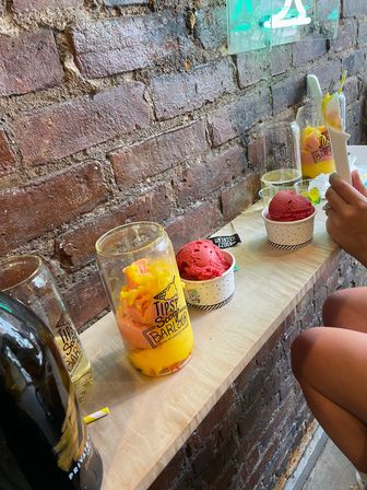 Colorful mango and raspberry sorbet scoops in cups and a tall jar on a narrow wooden counter against an exposed brick wall in an urban ice cream shop, a hand holding a spoon.