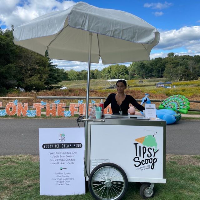 Woman vendor behind a white mobile boozy ice cream cart under a large umbrella at a sunny outdoor farm festival with fields and an inflatable peacock in the background.