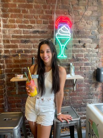 Smiling woman in white tank and shorts holding a colorful layered frozen drink with straws, sunglasses on her head, standing against an exposed brick wall under a neon ice cream sundae sign in a trendy parlor