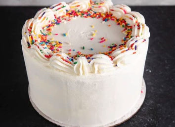 White frosted round cake with piped swirl border and colorful rainbow sprinkles on a dark countertop.