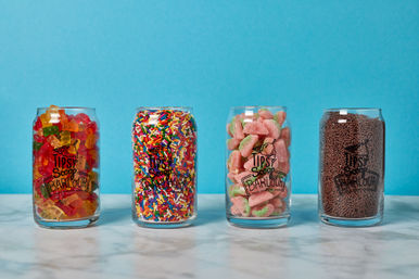 Four clear glass jars lined up on a marble surface against a bright blue background, each filled with colorful candy: gummy bears, rainbow sprinkles, pink-and-green sour watermelon slices, and chocolate sprinkles.