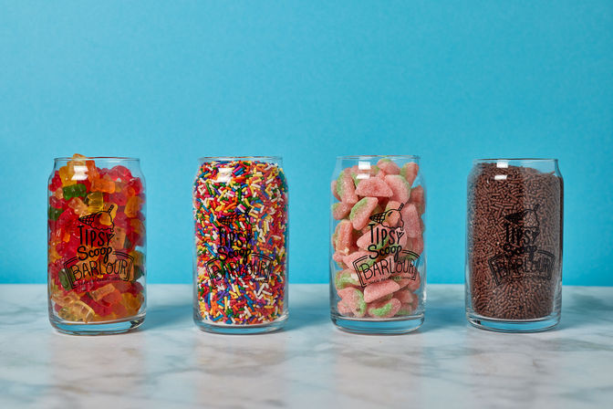 Four clear glass jars lined up on a marble surface against a bright blue background, each filled with colorful candy: gummy bears, rainbow sprinkles, pink-and-green sour watermelon slices, and chocolate sprinkles.