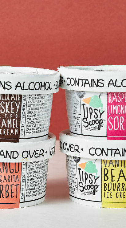 Playful stack of four alcohol-infused ice cream and sorbet pints—whiskey salted caramel, mango margarita, raspberry limoncello, vanilla bean bourbon—white lids reading 'contains alcohol' against a red backdrop