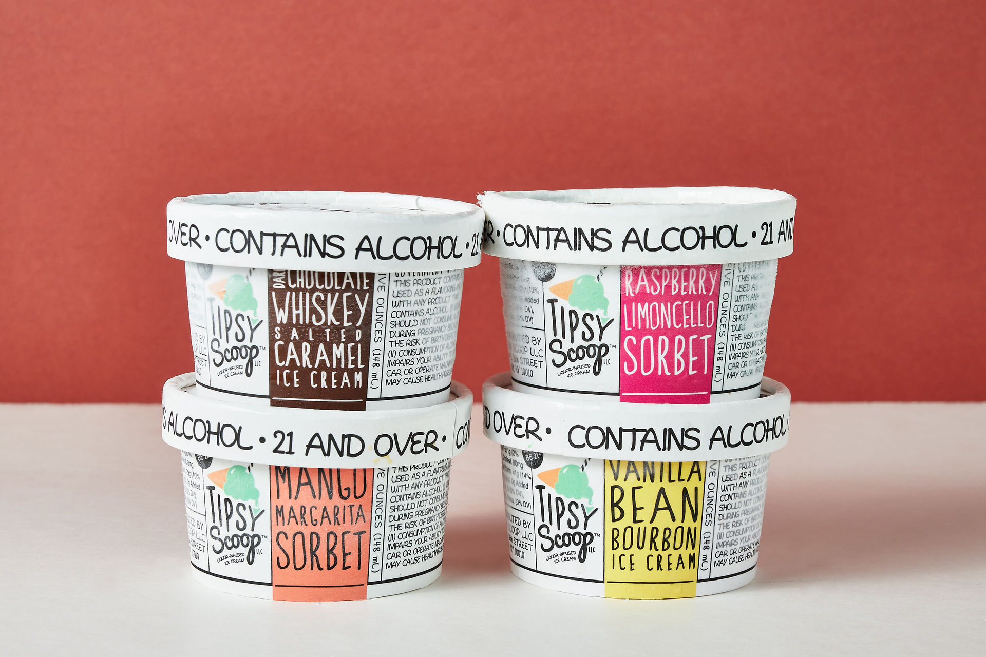 Playful stack of four alcohol-infused ice cream and sorbet pints—whiskey salted caramel, mango margarita, raspberry limoncello, vanilla bean bourbon—white lids reading 'contains alcohol' against a red backdrop
