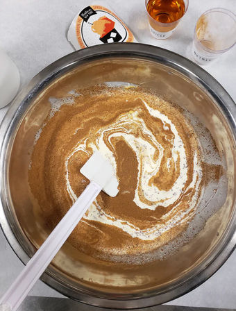 Stainless steel mixing bowl on a kitchen counter with creamy batter and pumpkin-spice swirl being stirred by a white spatula, spice jars and measuring cup nearby — cozy fall baking prep.