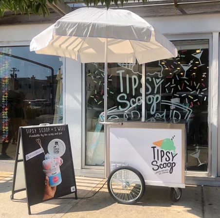 Outdoor mobile ice cream cart with a white umbrella parked on a sunny sidewalk outside a storefront with colorful sprinkle window art and a chalkboard sign showing a pastel swirled scoop.