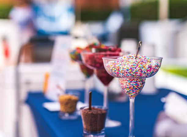 Festive rainbow sprinkles piled in a martini-style glass with a spoon on a blue table at an outdoor dessert toppings bar, with blurred bowls of chocolate crumbs and fruit in the background.