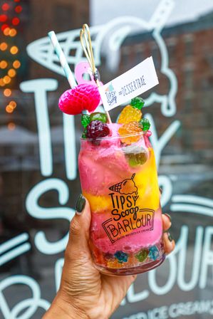 Hand holding a clear jar of colorful layered alcohol-infused ice cream dessert with bright pink and yellow swirls, gummy fruit candies, a striped paper straw and a cocktail pick with a small '#DESSERTBAE' flag, photographed in front of an ice cream shop window.