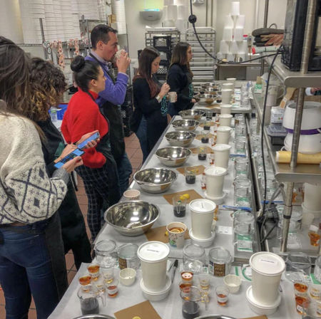 Group coffee cupping workshop in a roastery-style kitchen — participants sample brews at a long stainless-steel prep table lined with metal bowls, glass jars, small tasting cups and white to-go cups.