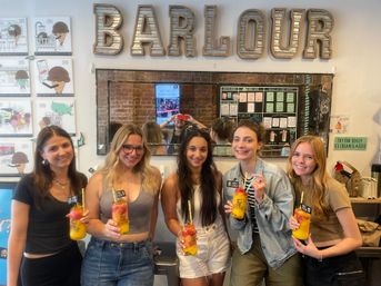 Five friends smiling inside a bright dessert shop, each holding a layered mango-and-berry boozy cocktail with straws, mirror and menu board visible behind them.
