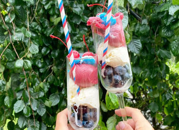 Two tall flutes held against a leafy garden backdrop filled with blueberries, vanilla ice cream, pink berry sorbet, cherries and festive red-white-blue striped straws — a refreshing summer dessert.