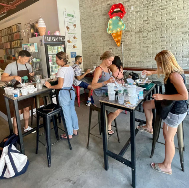 Group of people in aprons stirring tubs at a lively urban ice cream workshop with neon ice-cream cone sign and takeaway pints display