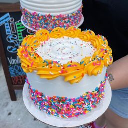 Colorful sprinkle-covered white layer cake with bright yellow buttercream rosettes on top, held outdoors near a chalkboard sign