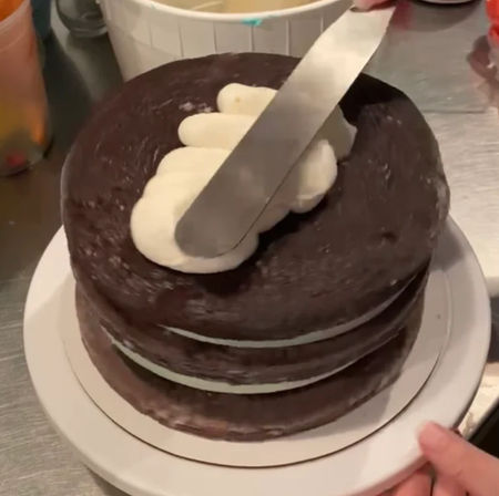 Three-layer chocolate cake on a turntable at a baking workspace, white buttercream between layers and being smoothed on top with an offset spatula
