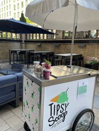 Urban outdoor patio with umbrella-shaded ice cream cart, stainless top holding colorful cups of liquor‑infused ice cream, lounge sofas and high-top tables in a city courtyard.