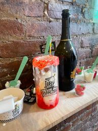 Liquor-infused milkshake in a tall glass with red sugar rim and melting vanilla ice cream, green spoons, gummy candies and a champagne bottle on a brick-wall dessert bar counter.