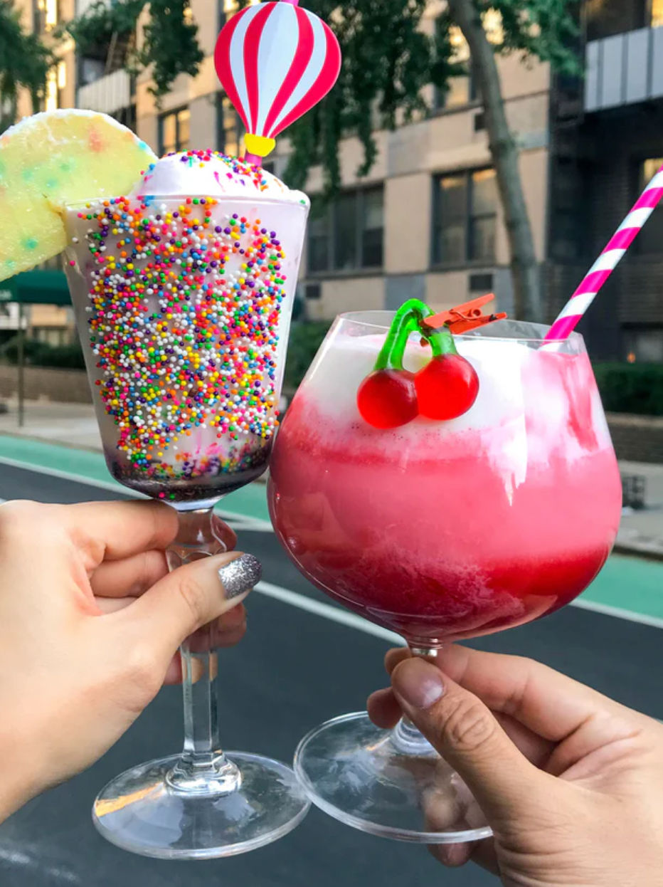 Two colorful dessert drinks clinking on an urban street — a sprinkle-covered milkshake with whipped cream, cookie wedge and hot‑air‑balloon topper, and a pink layered frothy cocktail with cherry candy garnish and striped straw.