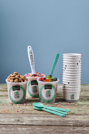 Party-ready dessert display: three paper cups with cookie pieces, rainbow sprinkles and gummy bears, turquoise spoons and stacked cups on a weathered wooden table against a blue backdrop.