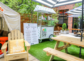 Sunny urban courtyard patio with a mobile ice cream cart, striped umbrellas, wooden picnic tables and an Adirondack chair outfitted with a "Hello Summer" pillow on artificial grass.