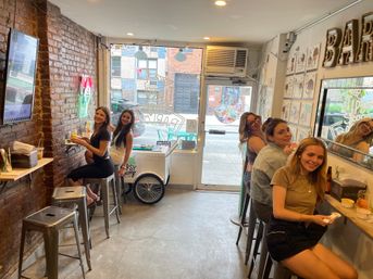 Group of friends enjoying ice cream at a cozy urban scoop shop with exposed brick wall, neon sign, metal bar stools and a street-facing window.