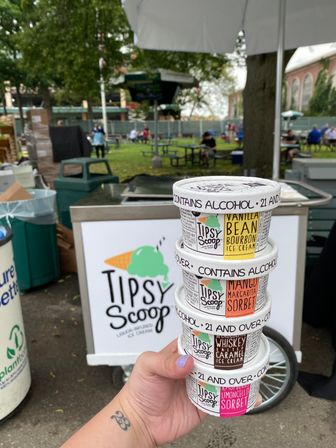 Hand holding a stack of four colorful alcohol‑infused ice cream cups in front of a park food cart at an outdoor summer festival, picnic tables and trees blurred in the background