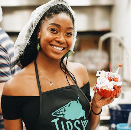 Smiling server wearing a hairnet and apron holding a colorful ice cream sundae with whipped cream, sprinkles and cherries in a waffle bowl at a dessert shop counter.
