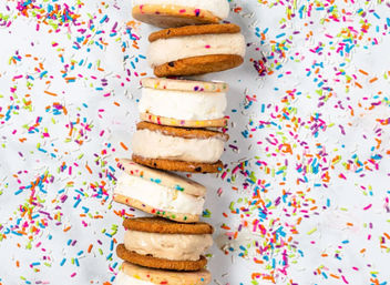 Stack of colorful ice cream cookie sandwiches on a white surface sprinkled with rainbow confetti sprinkles — fun party dessert close-up