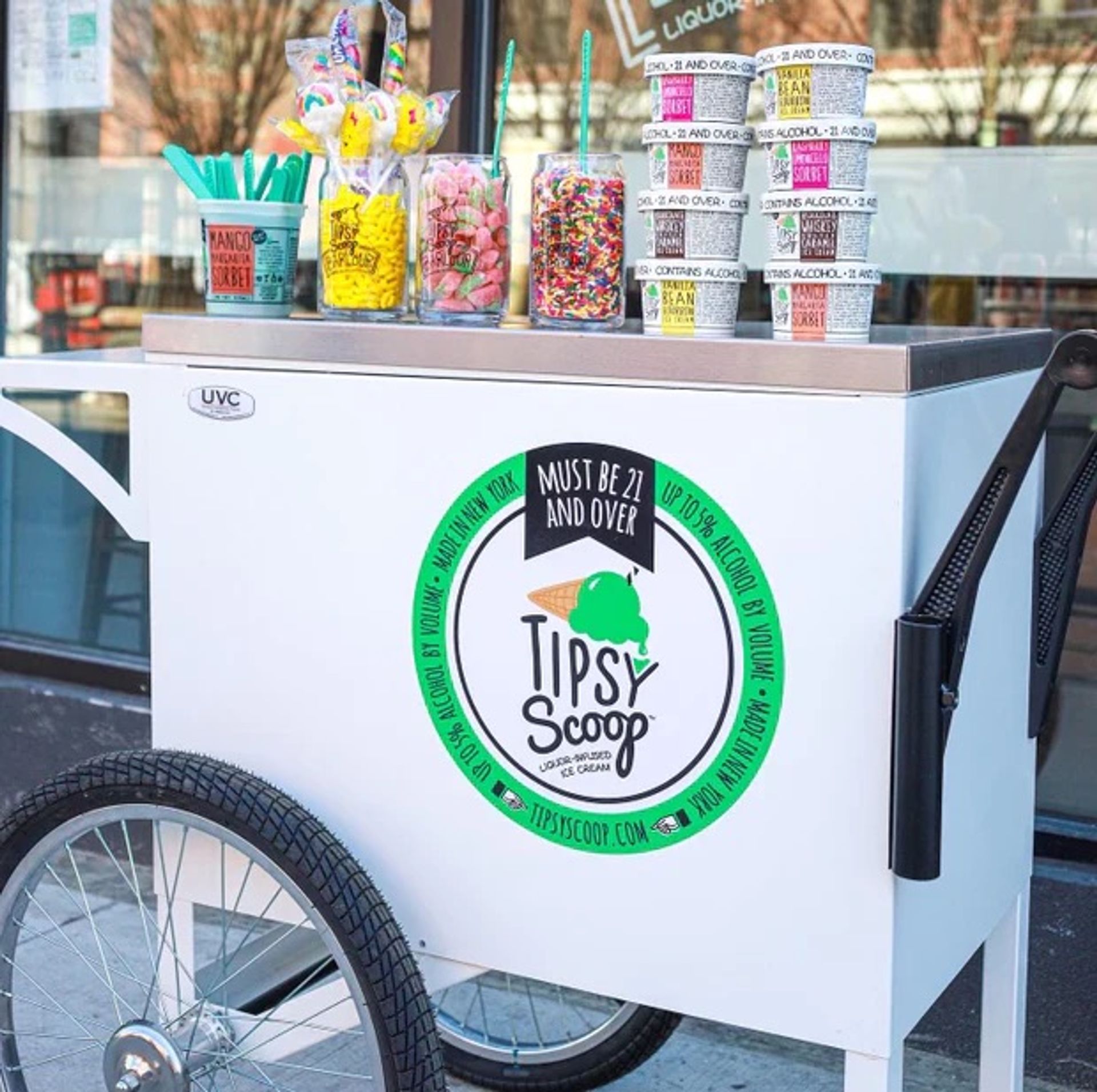 White mobile ice cream cart on a city sidewalk with a large wheel, jars of colorful candy and sprinkles, teal spoons and stacked paper cups, green circular logo and 'Must be 21 and over' label promoting alcohol-infused ice cream.