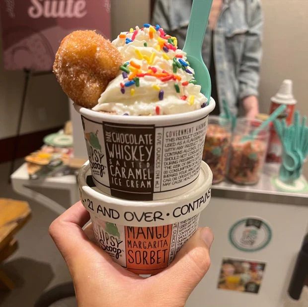 Hand holding stacked ice cream cups at a dessert counter — top cup topped with whipped cream, rainbow sprinkles, a cinnamon-sugar donut hole and a mint spoon; bottom cup with mango sorbet.