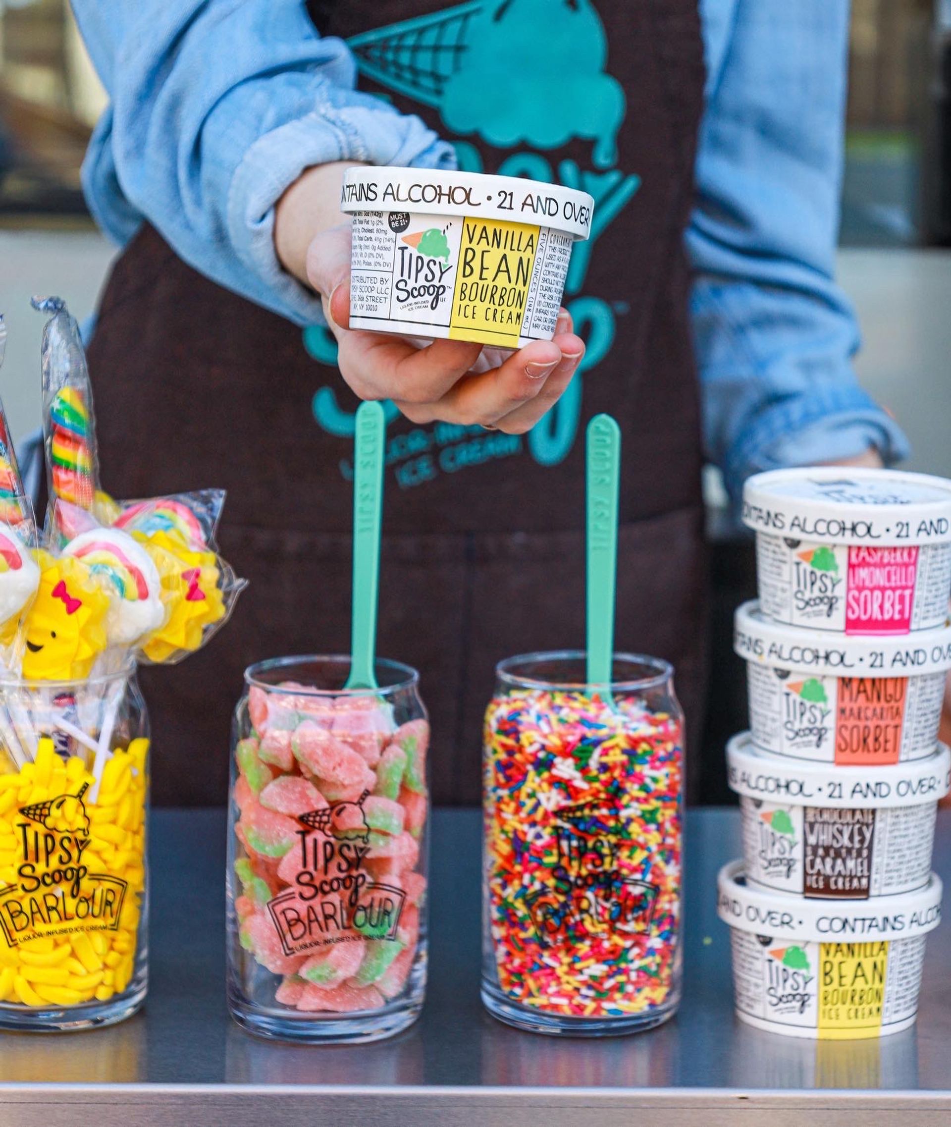 Hand presenting a mini boozy vanilla bean bourbon ice cream cup above jars of colorful candy and rainbow sprinkles at an outdoor ice cream stand.