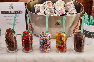 Event ice cream topping bar on a marble counter with five glass jars holding brownie chunks, red gummy cherries, rainbow sprinkles, gummy bears, and chocolate sprinkles, mint-green spoons, and a large metal tub of mini ice cream pints in the background.