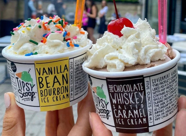 Close-up of two handheld cups of boozy ice cream — vanilla bean bourbon with whipped cream and colorful sprinkles, and chocolate whiskey salted caramel with whipped cream and a red cherry, outdoors on a city patio.
