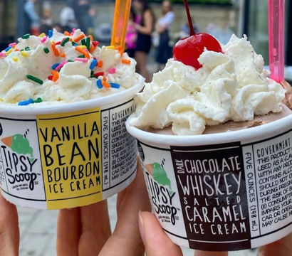 Two hands holding alcohol-infused ice cream cups outdoors: vanilla bean bourbon with rainbow sprinkles and chocolate whiskey salted caramel topped with whipped cream and a cherry.
