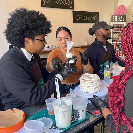 Cake-decorating workshop at a bakery counter, participants piping white icing onto a spiral layered cake with tools and tubs nearby.