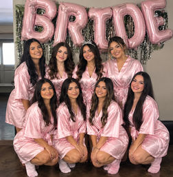 Bridal-party getting-ready photo of eight women in matching pink satin robes and fuzzy socks smiling indoors beneath large pink "BRIDE" balloons, bride wearing a "Bride" tiara.