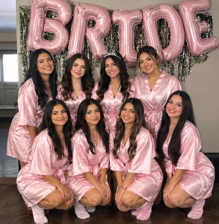 Bridal-party getting-ready photo of eight women in matching pink satin robes and fuzzy socks smiling indoors beneath large pink "BRIDE" balloons, bride wearing a "Bride" tiara.
