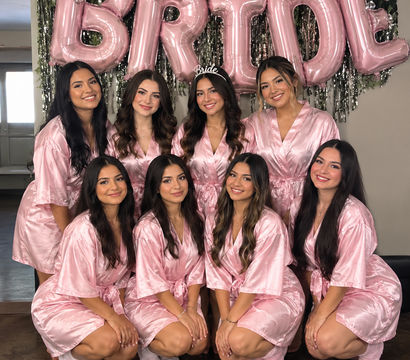 Bridal-party getting-ready photo of eight women in matching pink satin robes and fuzzy socks smiling indoors beneath large pink "BRIDE" balloons, bride wearing a "Bride" tiara.