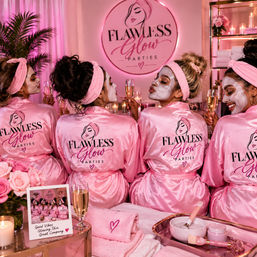 Four friends in matching pink satin robes and headbands wear white face masks and toast champagne at a pink-lit spa party surrounded by candles, roses and skincare bowls.