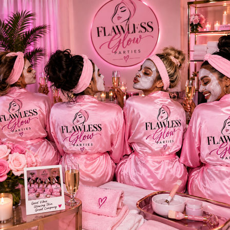 Four friends in matching pink satin robes and headbands wear white face masks and toast champagne at a pink-lit spa party surrounded by candles, roses and skincare bowls.