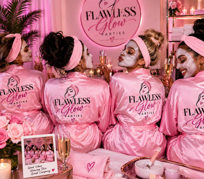 Four friends in matching pink satin robes and headbands wear white face masks and toast champagne at a pink-lit spa party surrounded by candles, roses and skincare bowls.