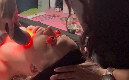 Spa scene of a client reclined receiving red LED light facial with protective eye shields while an esthetician glides a handheld device over the jawline, skincare tools and bowls on a nearby pink station.