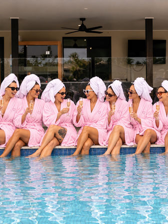 Group of women in pink robes and towel turbans, wearing sunglasses and laughing while clinking champagne glasses poolside at a resort