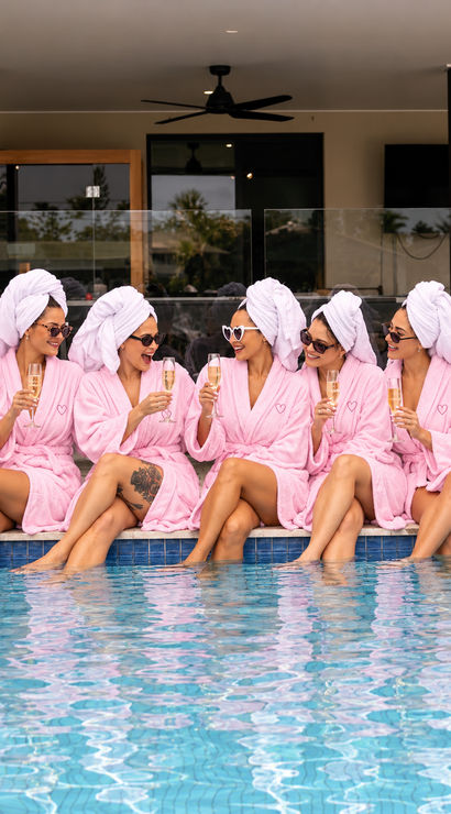 Group of women in pink robes and towel turbans, wearing sunglasses and laughing while clinking champagne glasses poolside at a resort