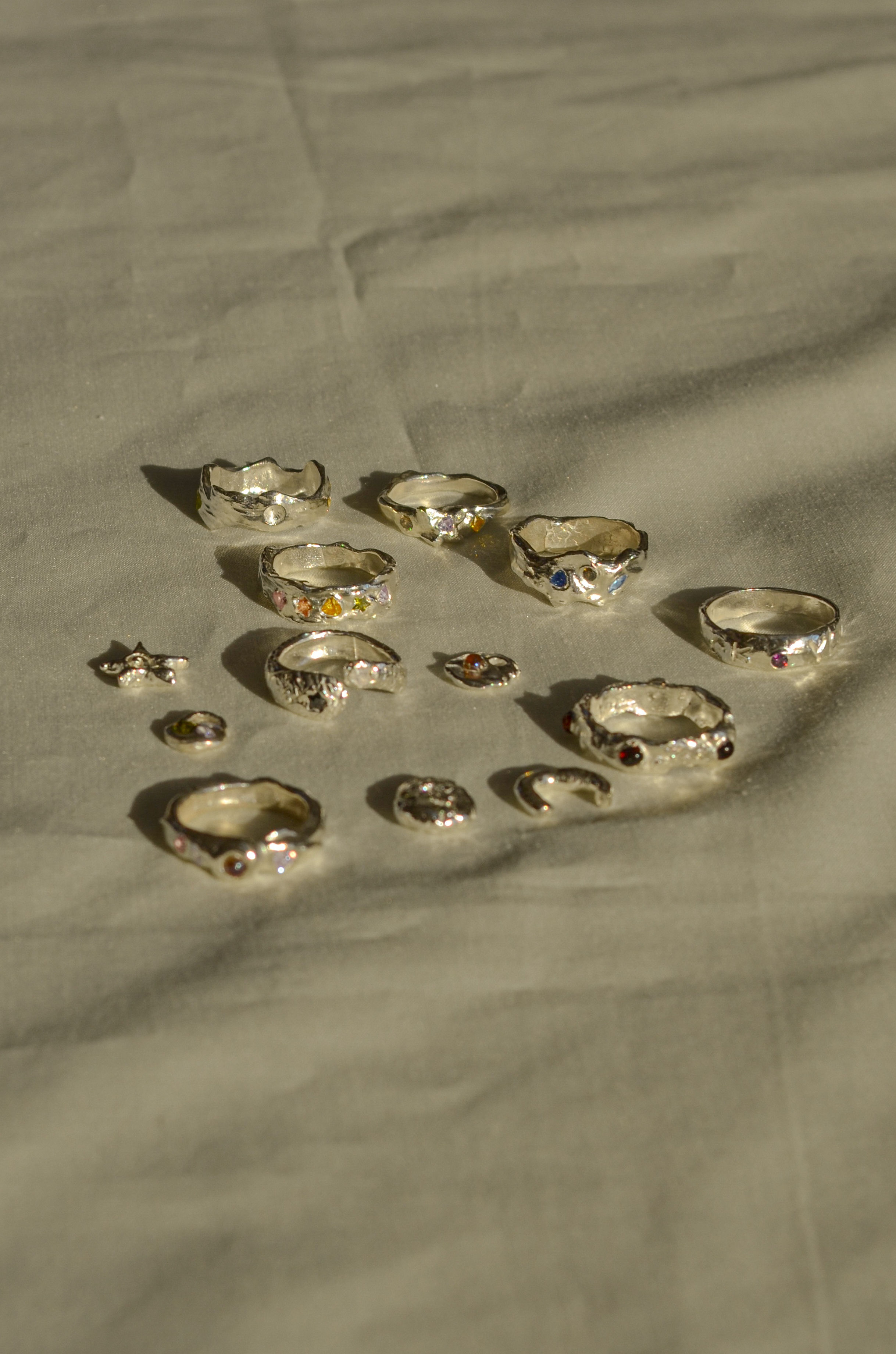 Sunlit flat-lay of assorted handmade silver rings and small charms with multicolored gemstones on soft beige fabric, casting warm shadows.