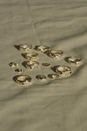 Sunlit flat-lay of assorted handmade silver rings and small charms with multicolored gemstones on soft beige fabric, casting warm shadows.