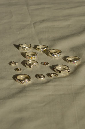 Sunlit flat-lay of assorted handmade silver rings and small charms with multicolored gemstones on soft beige fabric, casting warm shadows.