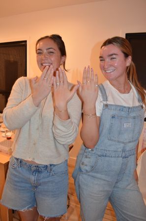 Two smiling friends in a home interior showing manicured hands with rings — one in denim overalls, the other in a cardigan and denim shorts.